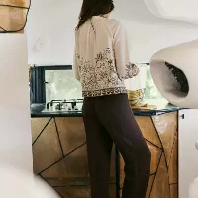 Pessoa de pé numa cozinha com blusa creme bordada e calças castanhas