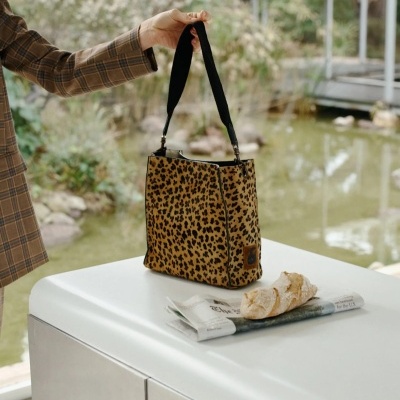 Mala animal print castanha e preta com alça preta, segurada por pessoa, sobre mesa branca com pão e jornal