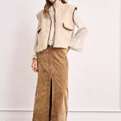 Mulher vestindo colete de pêlo creme, saia castanha longa e blusa branca com detalhes em renda