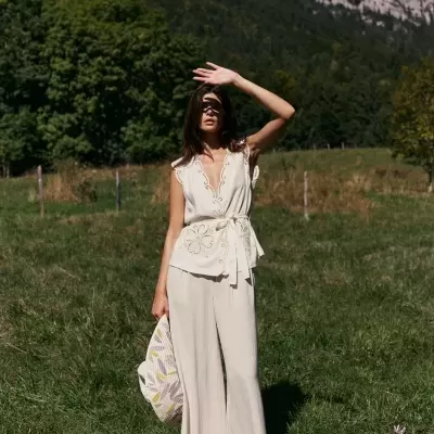 Mulher com conjunto branco com bordados florais e calças largas, segurando saco têxtil, num campo verde com montanhas ao fundo