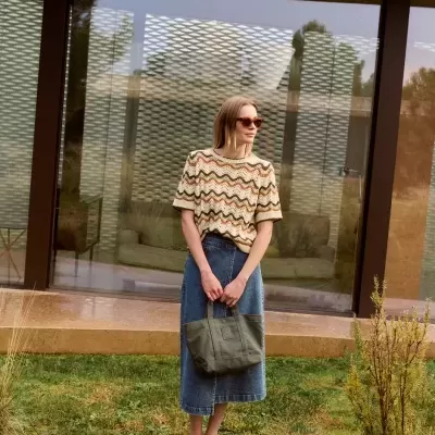 Mulher com camisola de malha bege com padrão ondulado, saia midi de ganga azul e bolsa verde numa área exterior