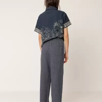 Pessoa de costas com camisa azul escura com desenho floral bege, calças cinza riscas, sandálias azuis