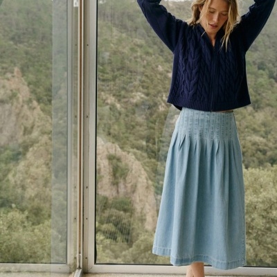 Mulher com camisola de malha azul escura e saia comprida azul clara perto de janela grande
