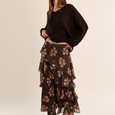 Mulher com camisola castanha escura, saia floral com folhos e botas castanhas em fundo neutro