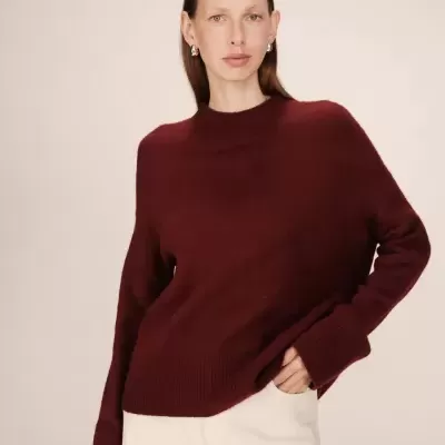 Mulher com camisola bordô de malha e calças brancas em fundo bege.