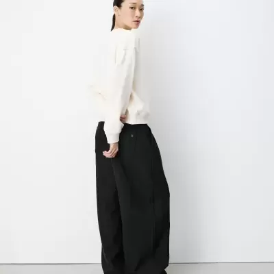 Pessoa de cabelo preso com camisola branca e calças largas pretas de costas num fundo branco.