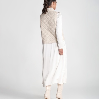 Mulher de costas com colete bege acolchoado, vestido branco e botas bege de salto alto.