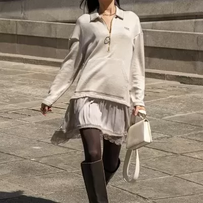 Mulher com vestido bege, botas pretas e mala bege a caminhar ao ar livre em calçada de pedra.