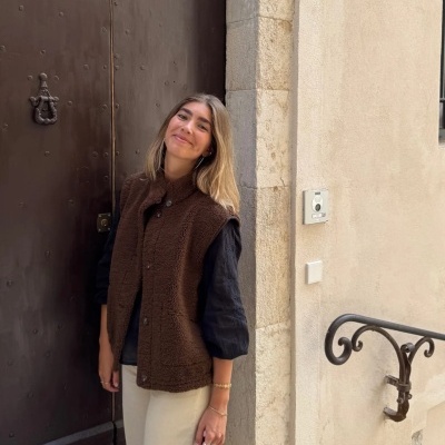 Mulher com colete castanho e camisa preta junto a porta de ferro castanha