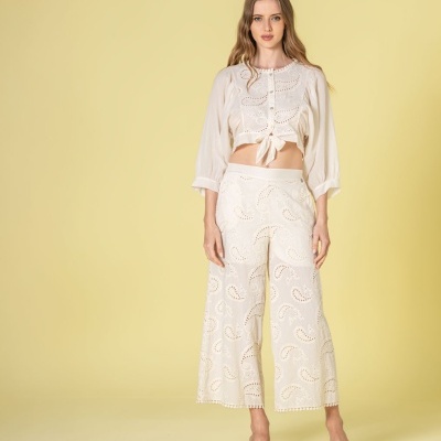 Modelo veste conjunto feminino creme com camisa crop e calças largas bordadas, sandálias brancas, fundo amarelo.