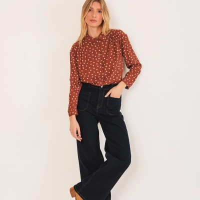 Mulher com camisa castanha de corações brancos, calças pretas largas e botas castanhas em fundo branco.