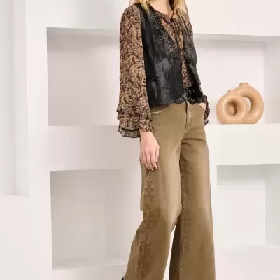 Mulher com blusa castanha, colete preto, calças largas castanhas e botas pretas em ambiente branco minimalista.