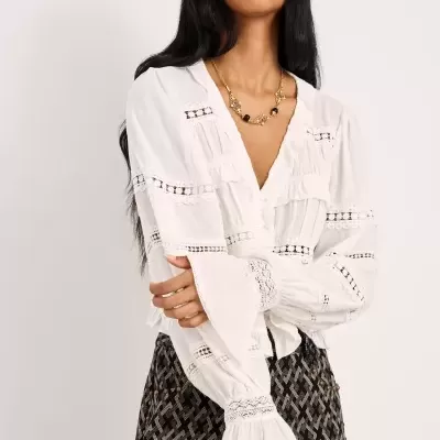 Mulher a usar camisa branca com renda e calças com padrão geométrico escuras, colar dourado com pedras, fundo branco.