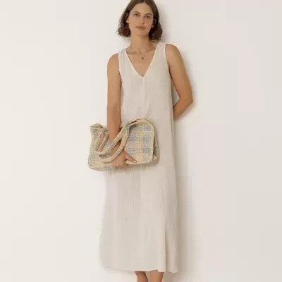Mulher com vestido branco longo sem mangas segurando bolsa trançada azul e bege e calçando sapatos brancos.