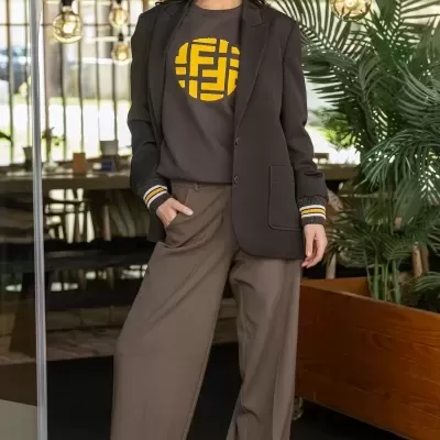 Mulher com blazer castanho escuro, camisola castanha com padrão amarelo, calças largas castanhas, sapatos castanhos claros.