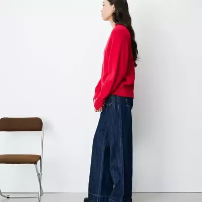 Mulher de perfil com camisola vermelha e calças de ganga largas em fundo branco próximo de cadeira marrom
