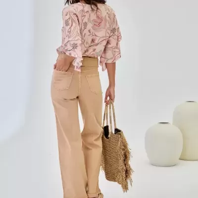 Mulher com blusa floral rosa, calças bege largas e bolsa de palha com franjas