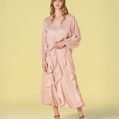 Mulher com blusa e saia rosa claro com mangas bufantes e detalhes franzidos, sandálias castanhas, fundo amarelo.