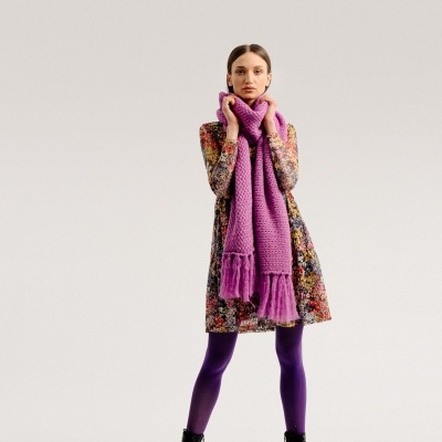 Pessoa a vestir vestido estampado de flores, cachecol rosa, collants roxos e botins pretos.