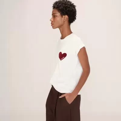 Mulher de perfil com t-shirt branca com coração vermelho e calças castanhas em fundo neutro