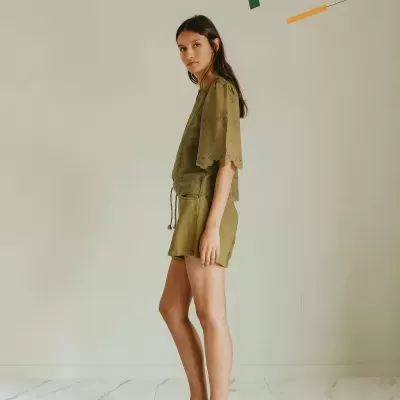 Mulher de pé com túnica e calções castanhos claros e sandálias cinzentas em ambiente claro com piso marmoreado.