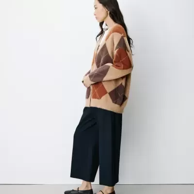Mulher com cardigan argyle e calças pretas de lado em fundo branco