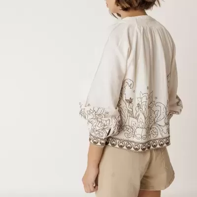 Blusa branca com padrão floral castanho na parte inferior e mangas, usada com calções bege.