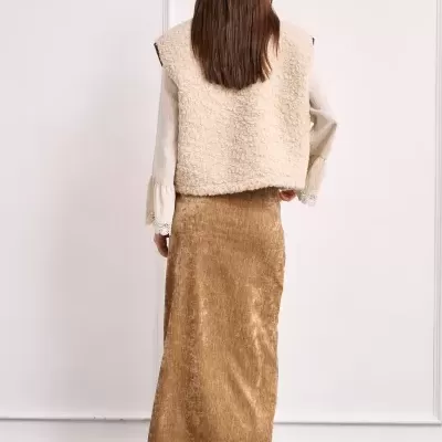 Mulher vista de costas a usar colete de pelo bege, blusa creme, saia castanha de veludo e botas castanhas.