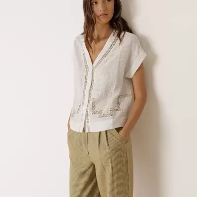 Mulher com blusa branca bordada e calças largas bege às riscas encostada à parede