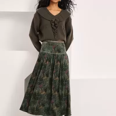 Mulher com camisola verde escura e saia estampada verde com botas pretas em fundo branco.