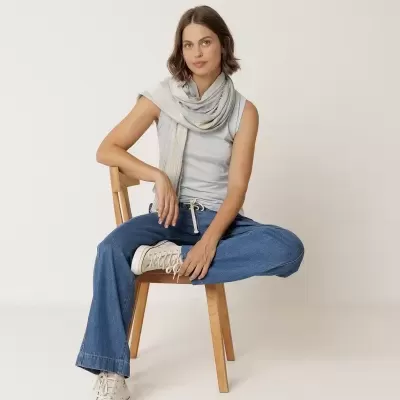 Mulher com camisa branca sem mangas, calças de ganga, sapatilhas e lenço bege claro sentada numa cadeira de madeira