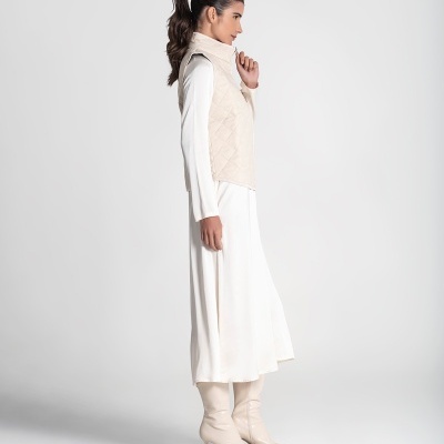 Mulher de vestido branco, colete bege acolchoado e botas bege de salto.