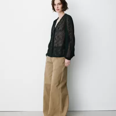 Mulher vestindo blusa preta de renda transparente com padrão floral e calças largas bege.