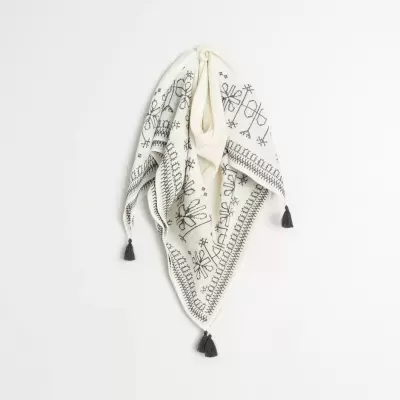 Pano triangular branco com bordados pretos e franjas pretas pendurado na parede