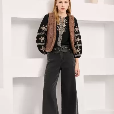 Look feminino com túnica preta bordada, colete castanho de pelo e calças pretas largas em fundo branco