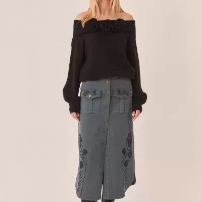 Mulher com chapéu preto a usar camisola preta ombro a ombro com flores e saia azul bordada com botões e botas pretas.