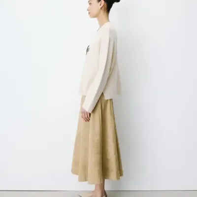 Mulher com camisola bege, saia midi bege e sapatos rasos cor bege junto a uma parede branca