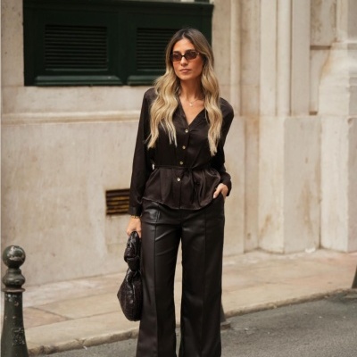 Mulher vestida de preto com blusa acetinada, calças de couro, sapatos de salto e mala, numa rua urbana.