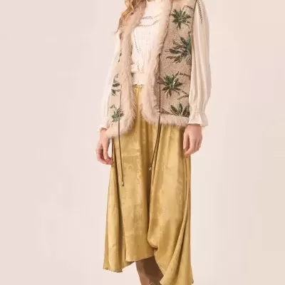 Mulher com casaco bege bordado, blusa branca, saia amarela e botas castanhas