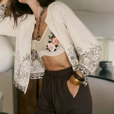 Mulher com top de crochet florido, casaco bege estampado e calças castanhas em ambiente interior