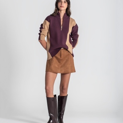 Mulher com sweater de malha bege e bordeaux, saia castanha curta e botas castanhas altas em fundo cinza.