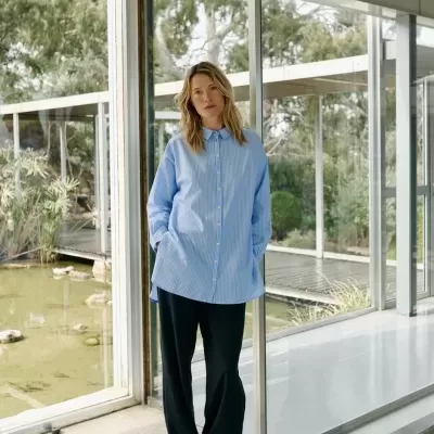 Mulher com camisa azul de algodão às riscas, calças pretas e sapatilhas castanhas ao lado de janelas de vidro