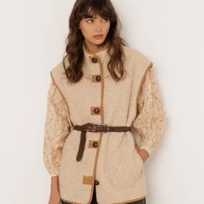 Mulher vestindo colete bege sherpa com botões e cinto, camisa floral pastel e calças pretas
