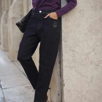 Mulher vestindo polo roxo de manga comprida, calças de ganga preta, sapatos de salto castanhos, mala preta trançada, encostada a parede de pedra