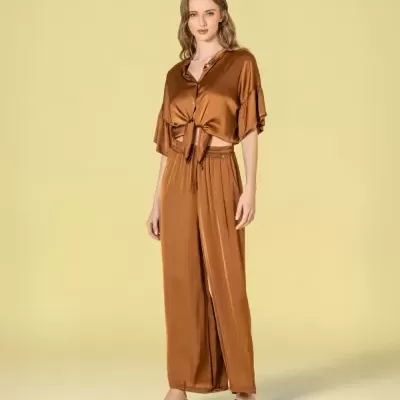 Modelo veste conjunto acetinado castanho, camisa e calças largas, calça sandálias pretas, fundo amarelo claro.