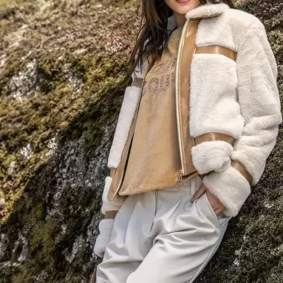 Mulher com casaco de pele sintética creme e bege e calças largas bege claro encostada em muro coberto de musgo