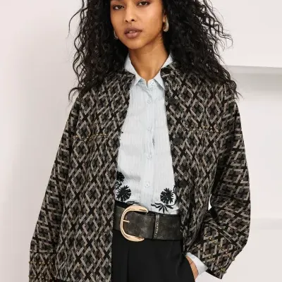 Mulher de cabelo cacheado com casaco preto e castanho, camisa azul e calças pretas com cinto.