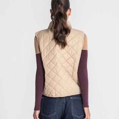 Colete acolchoado bege visto de costas com sweater de mangas compridas em bege e vinho e calças de ganga azul.