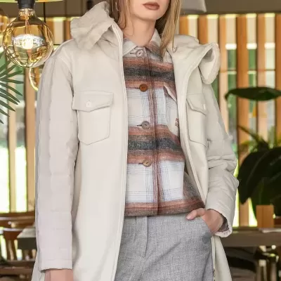 Modelo feminino vestindo casaco bege com gola de pelo, camisa xadrez castanha e cinzenta, e calças cinzentas.