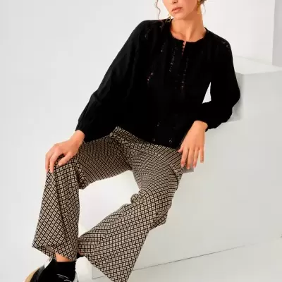 Mulher sentada com calças largas estampadas, blusa preta e sapatos pretos em fundo branco.